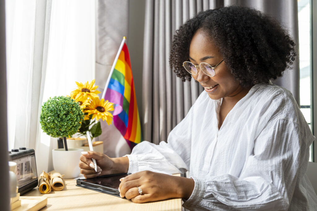 A photograph of a black woman and LGBT+ ally sat at a desk, with an LGBT+ rainbow pride flag.