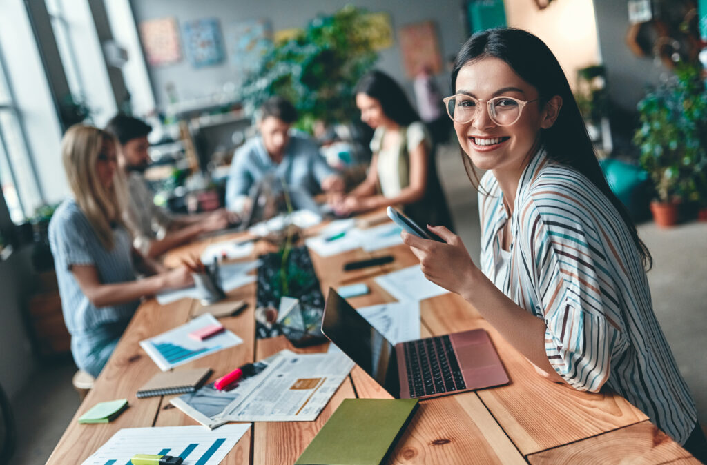 A photo of a young woman sat at work on her laptop, with her colleagues in the background.